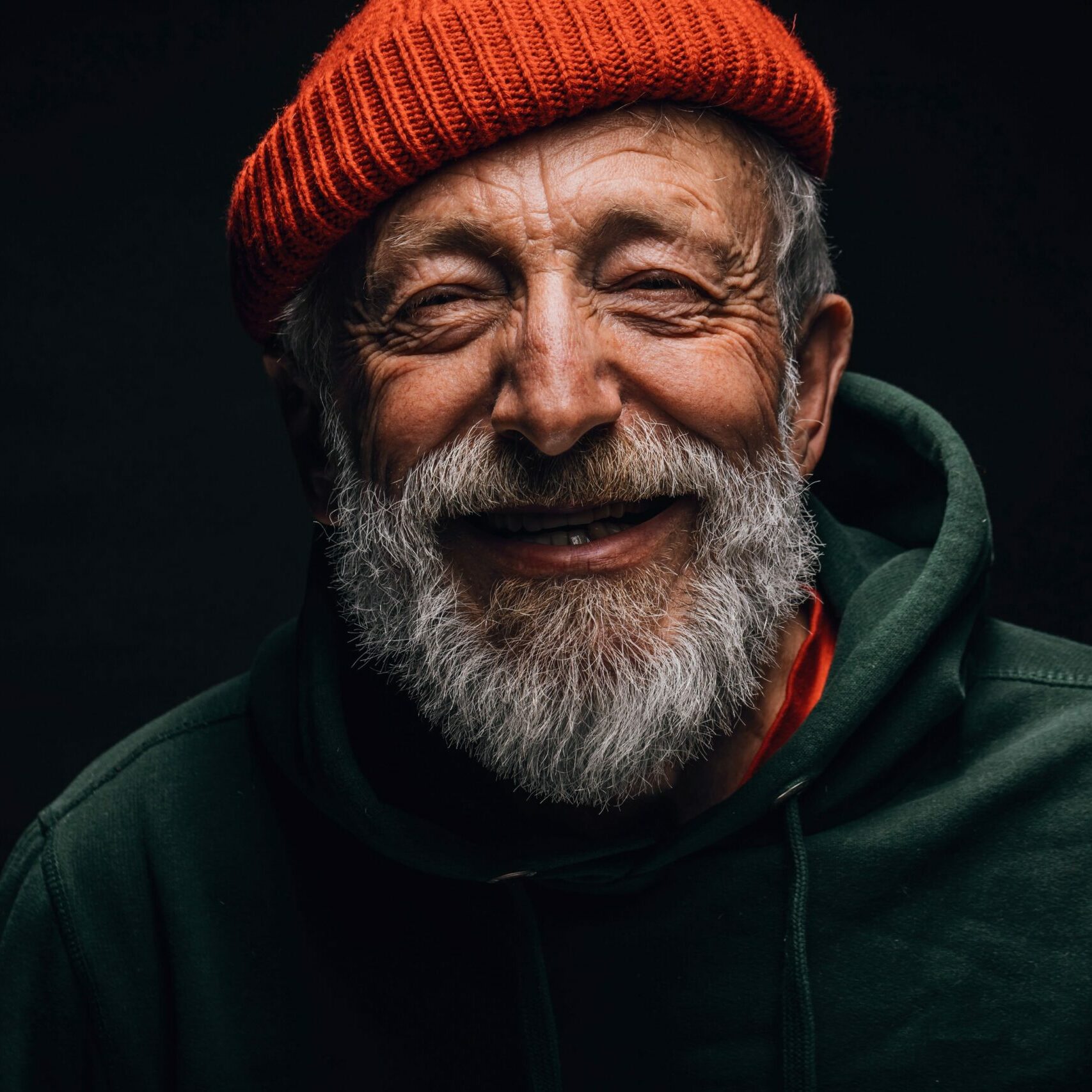 Ein alter, freundlich lächelnder Herr mit grauem Bart, einer roten Woll-Strickmütze und dunkelgrünemHoodie.